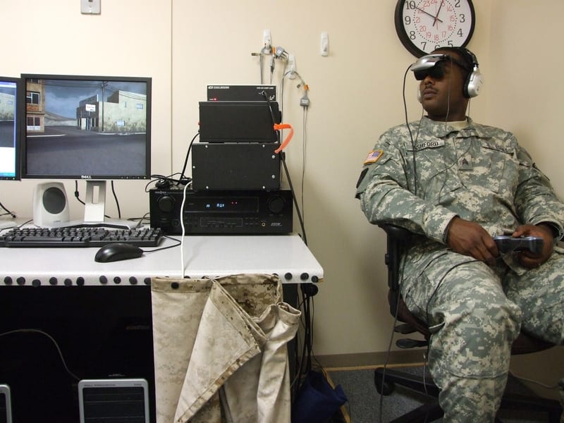 soldier sitting on a chair in a testing room wearing virtual reality headset next to a screen and tech equipment